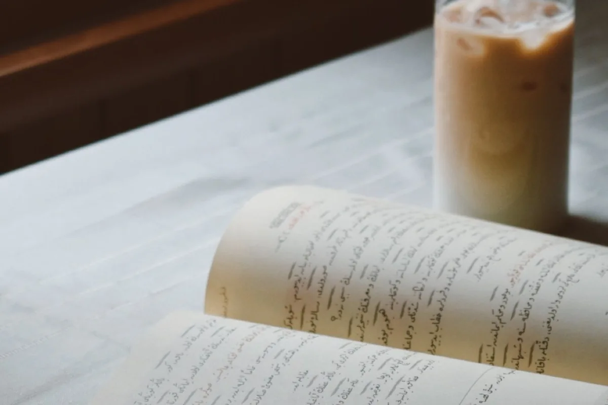 An open book and a glass of iced coffee on a table by a window